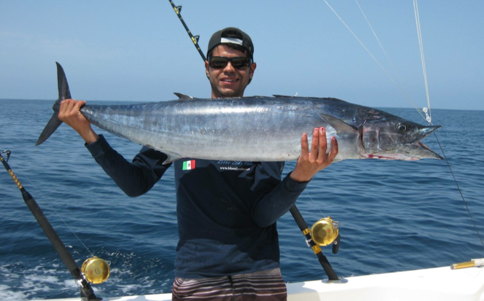 Man in sunglasses and baseball cap fishing from a boat, holding a large fish over the ocean.