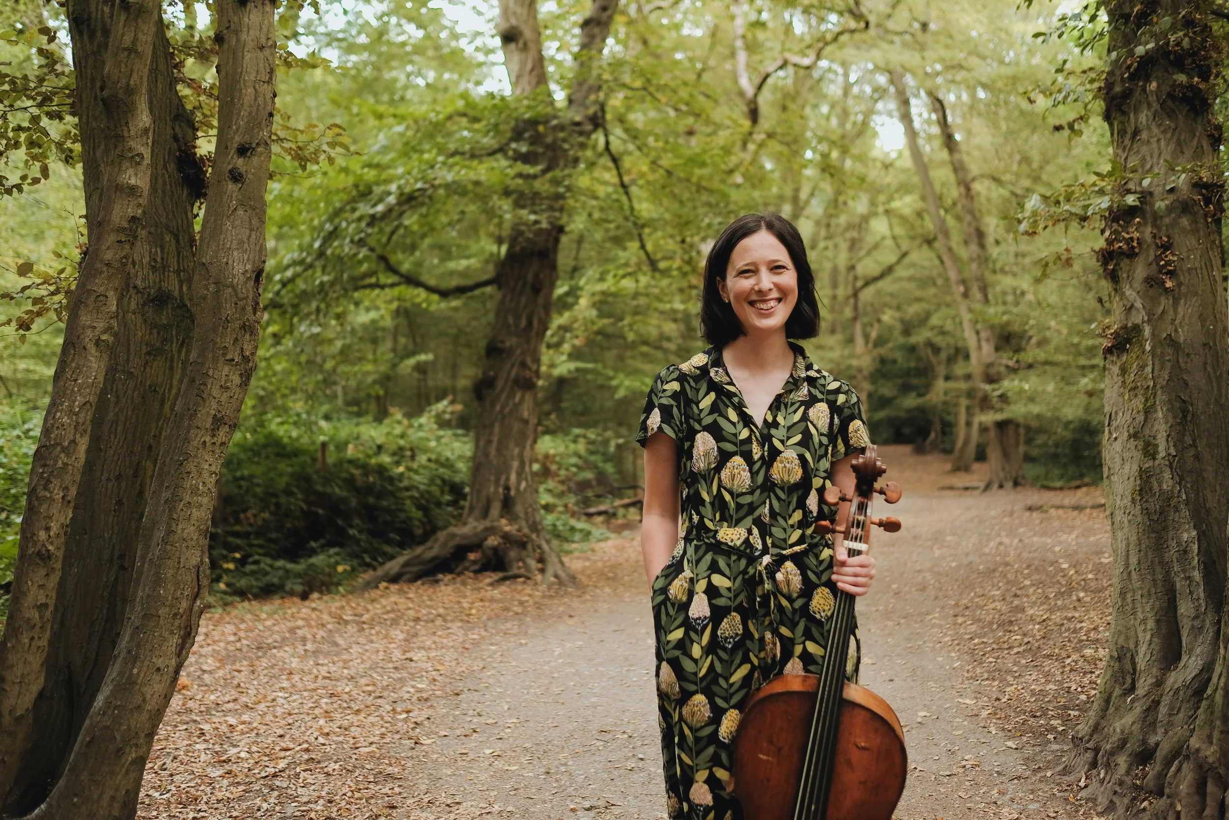 A woman with dark hair smiling while holding a cello stands among trees on a dirt trail in a forest.