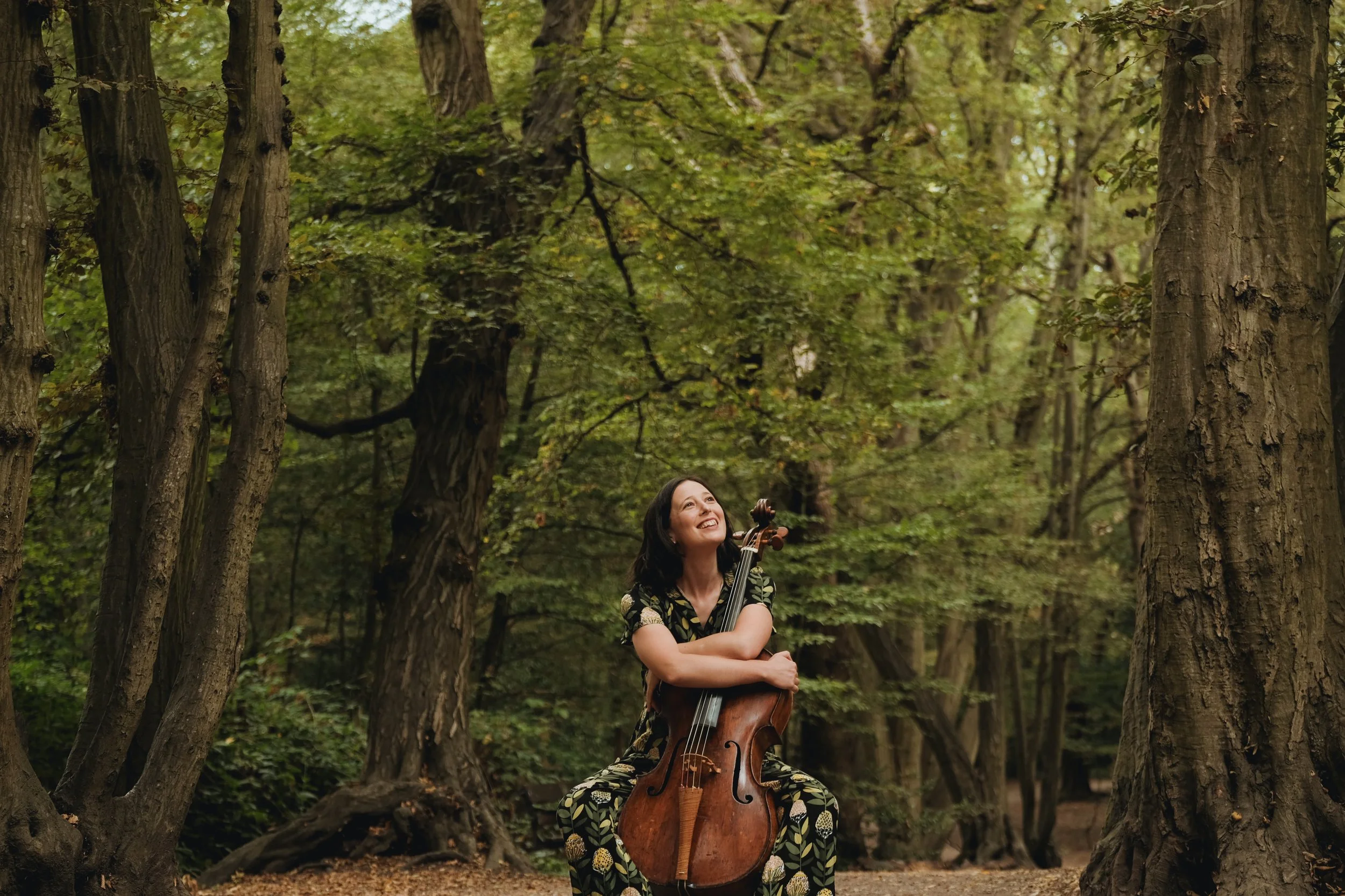 A woman sitting outdoors in a forest, holding and playing a cello, looking happy while surrounded by tall trees with green leaves.