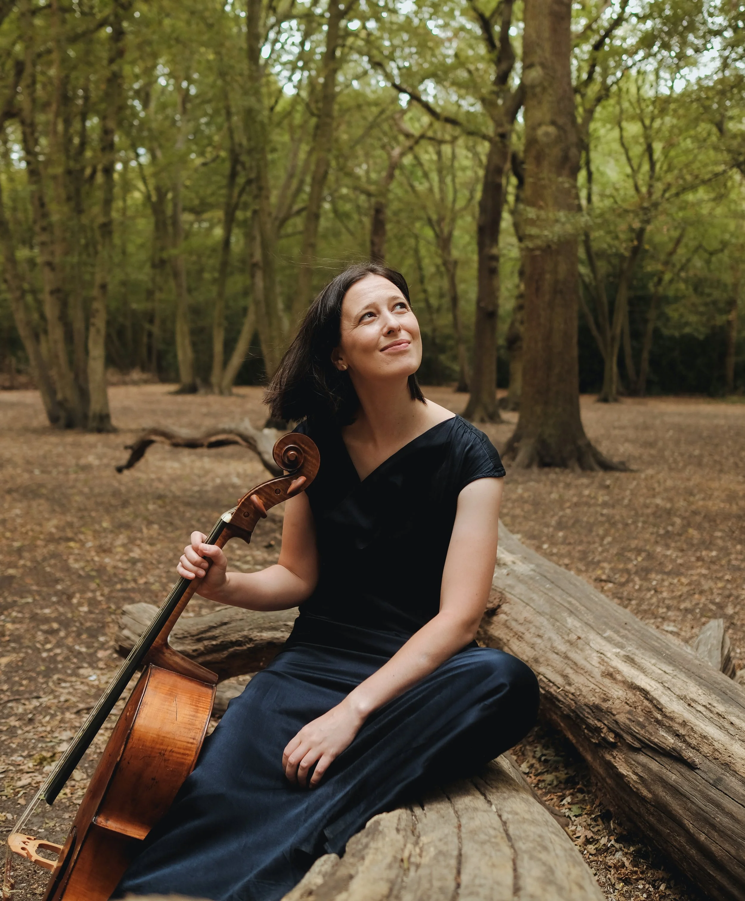 A woman with dark hair sitting on a fallen log in a forest, holding a guitar, and looking up with a smile among tall trees with green foliage.