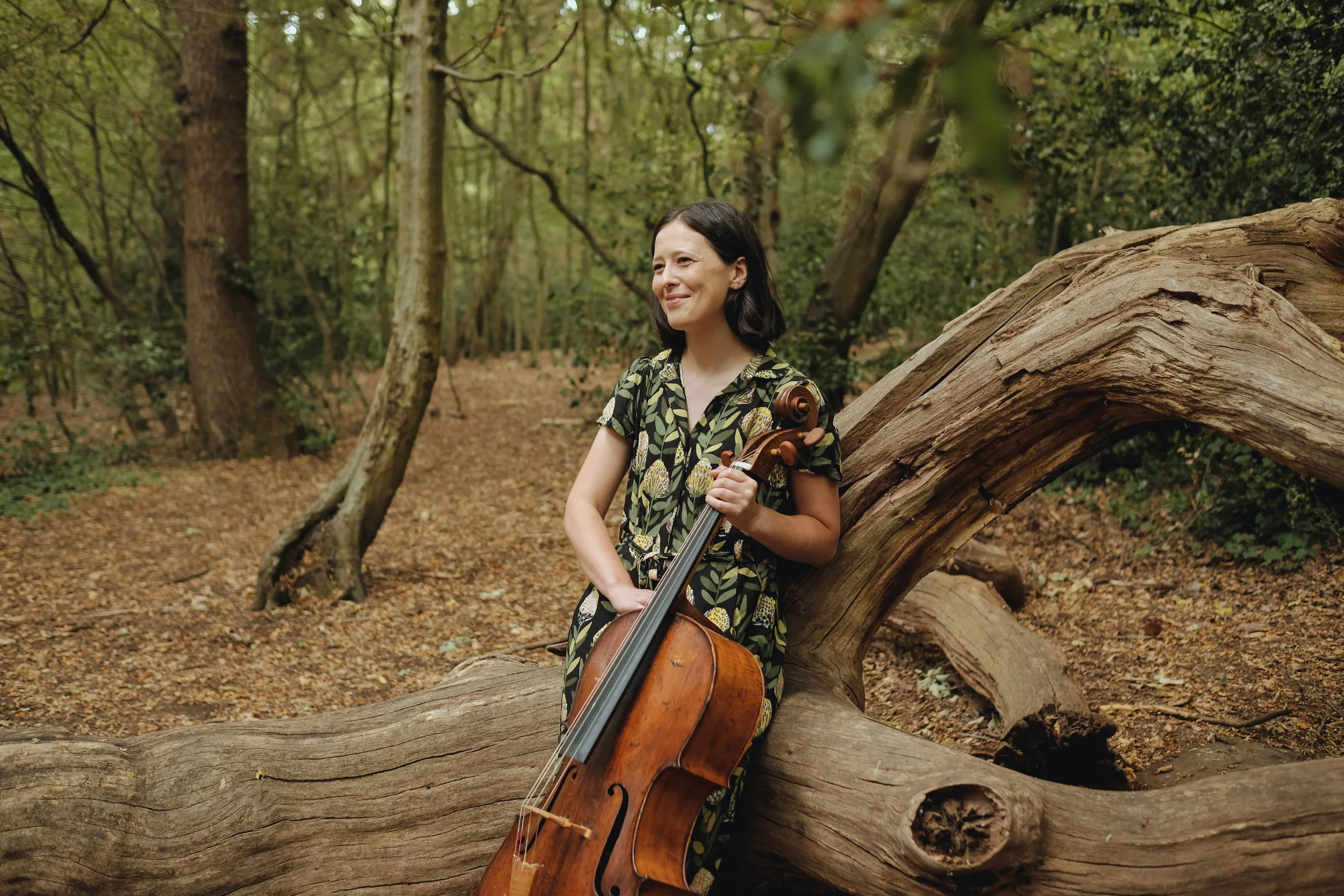 A woman stands outdoors in a forest, holding a cello and leaning against a fallen tree. She is smiling and looking to her left, wearing a short-sleeved, patterned dress with green and yellow leaves.
