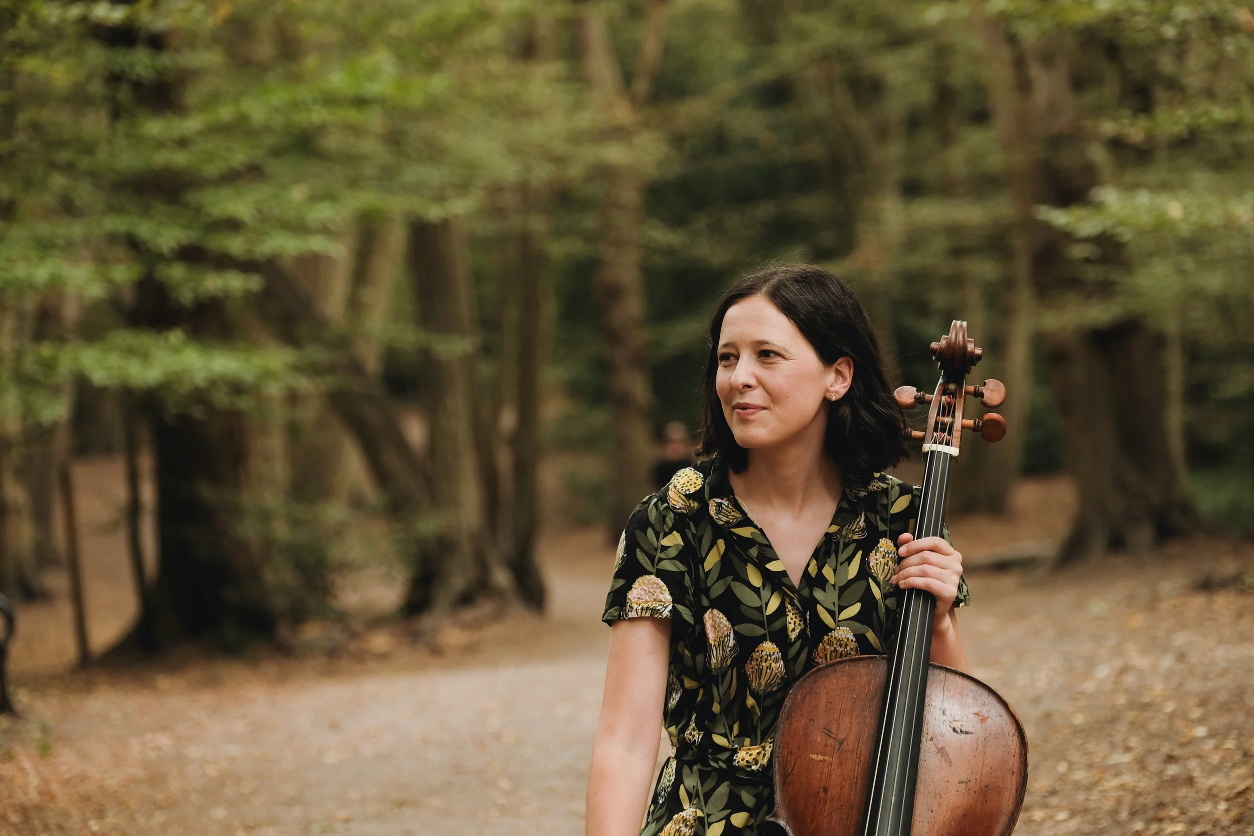 A woman with dark hair holding a cello outdoors in a park with trees.
