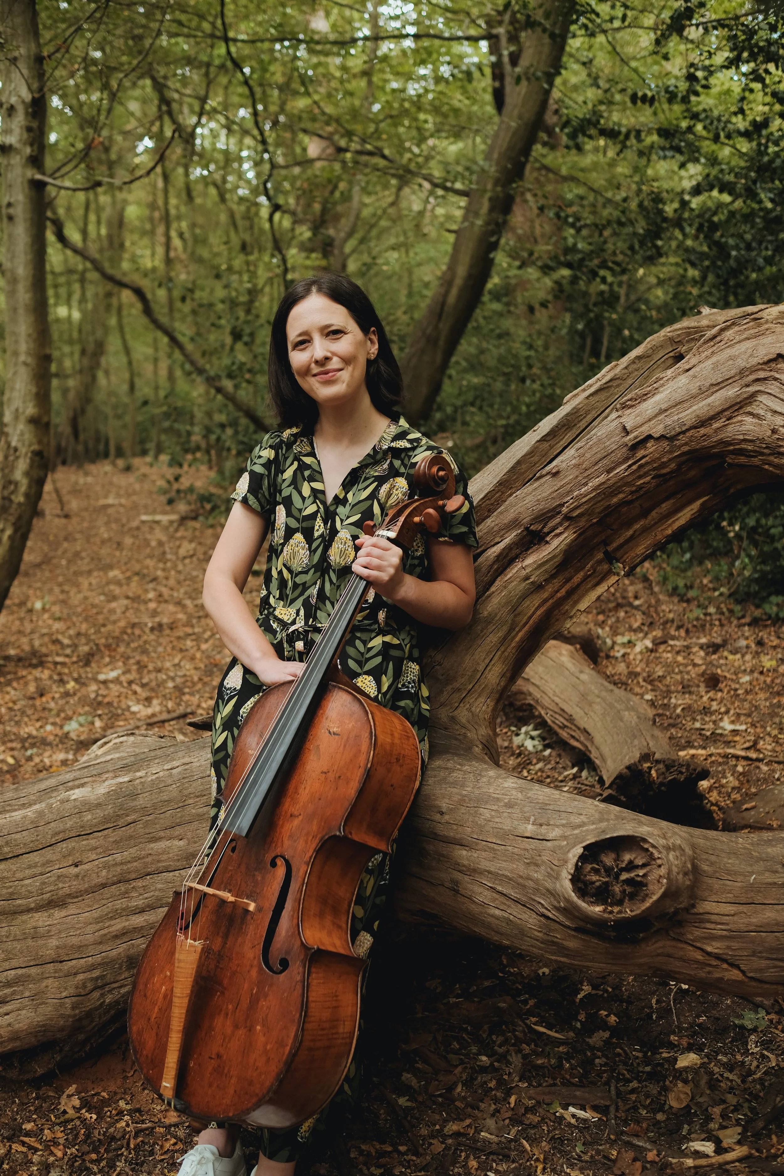 A woman with dark hair smiling while holding a cello is standing in a wooded area with trees and fallen leaves, leaning against a fallen tree trunk.