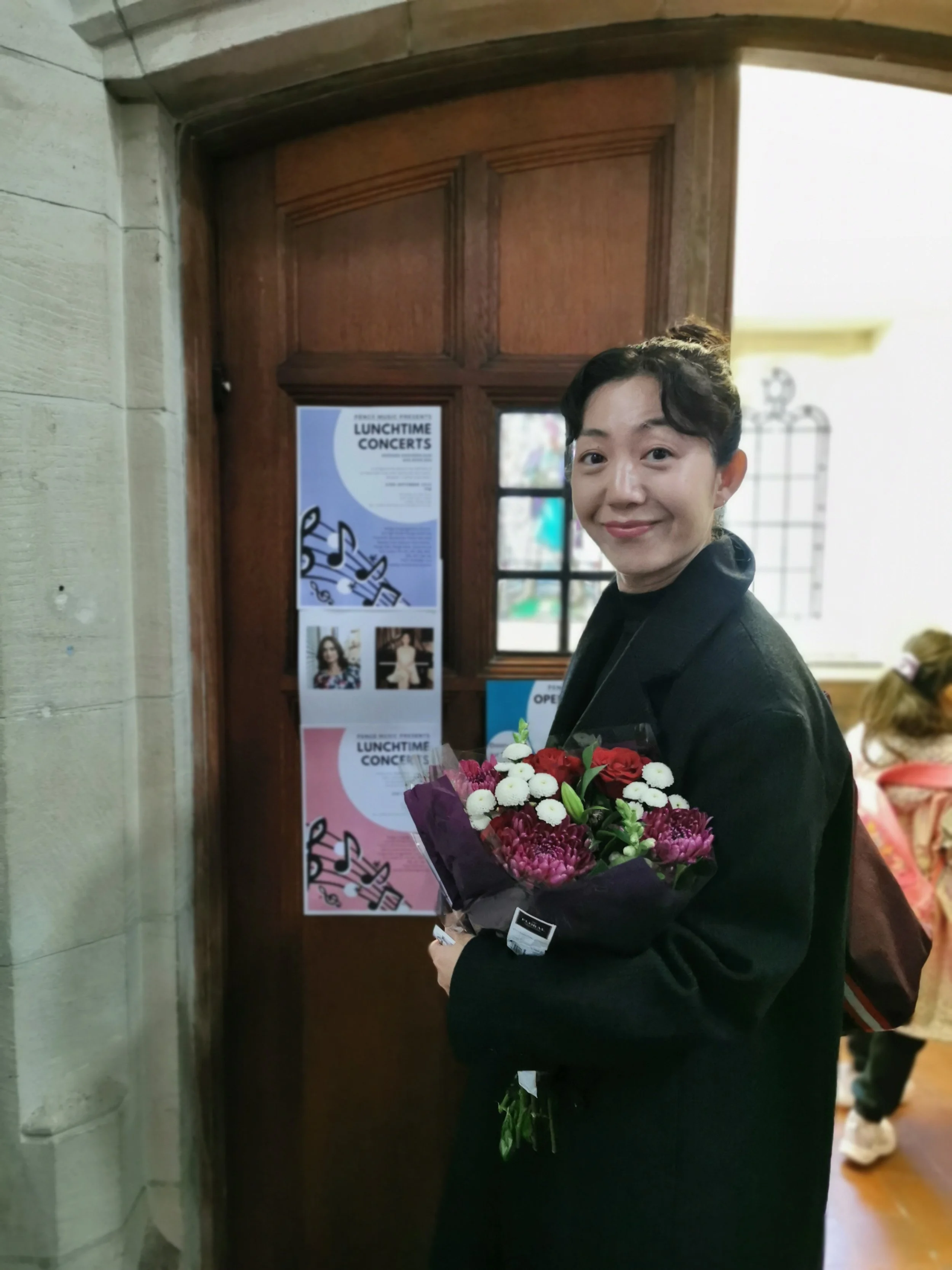 Smiling person in black clothing holding a colorful bouquet of flowers in front of a wooden door with a lunchtime concert poster.
