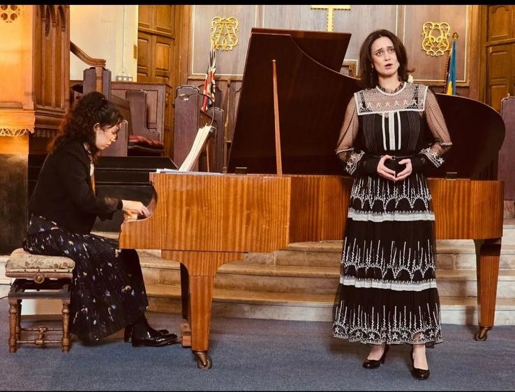 Female singer performing beside a grand piano while a pianist plays in an ornate church interior.
