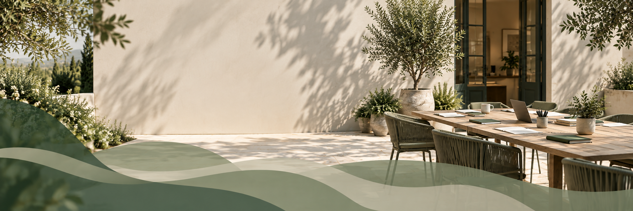 Outdoor patio with a wooden table, laptops, notebooks, potted plants, and chairs, nearby a white wall with potted trees and plants, sunlight creating shadows.