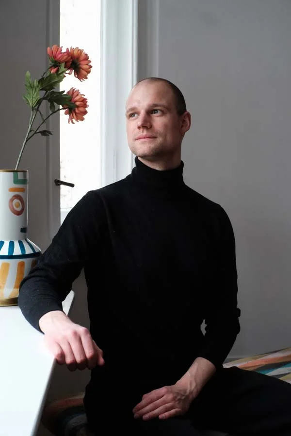 Thomas, Beziehungsberater bei connected., sitzt im schwarzen Rollkragenpullover neben einem Fenster mit Blumenstrauß