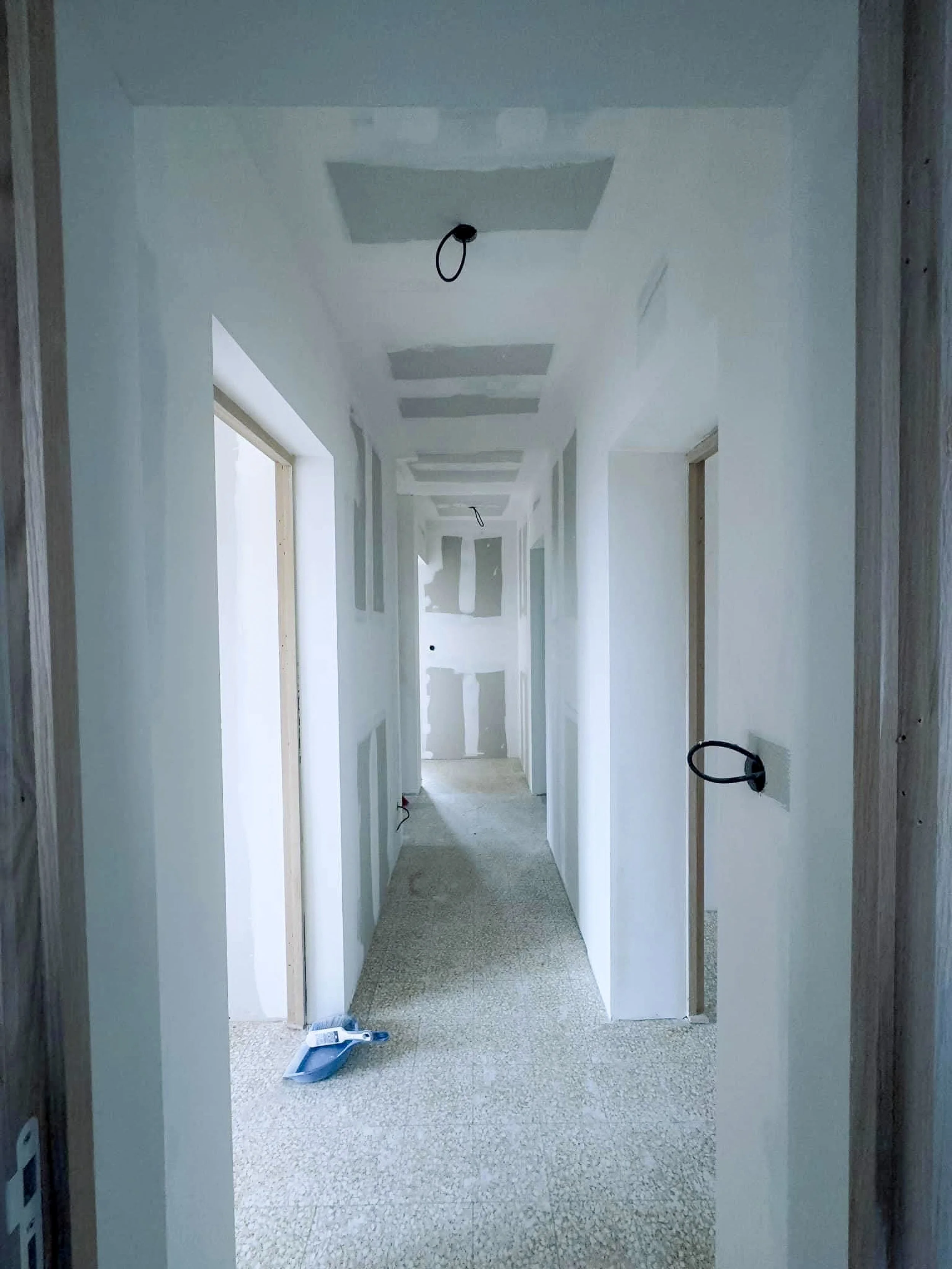 Un couloir en cours de construction avec des murs peints en blanc, des plafonds avec des structures en béton, des ouvertures de portes sans portes encore installées, des câbles électriques suspendus au plafond, et des outils de construction au sol.