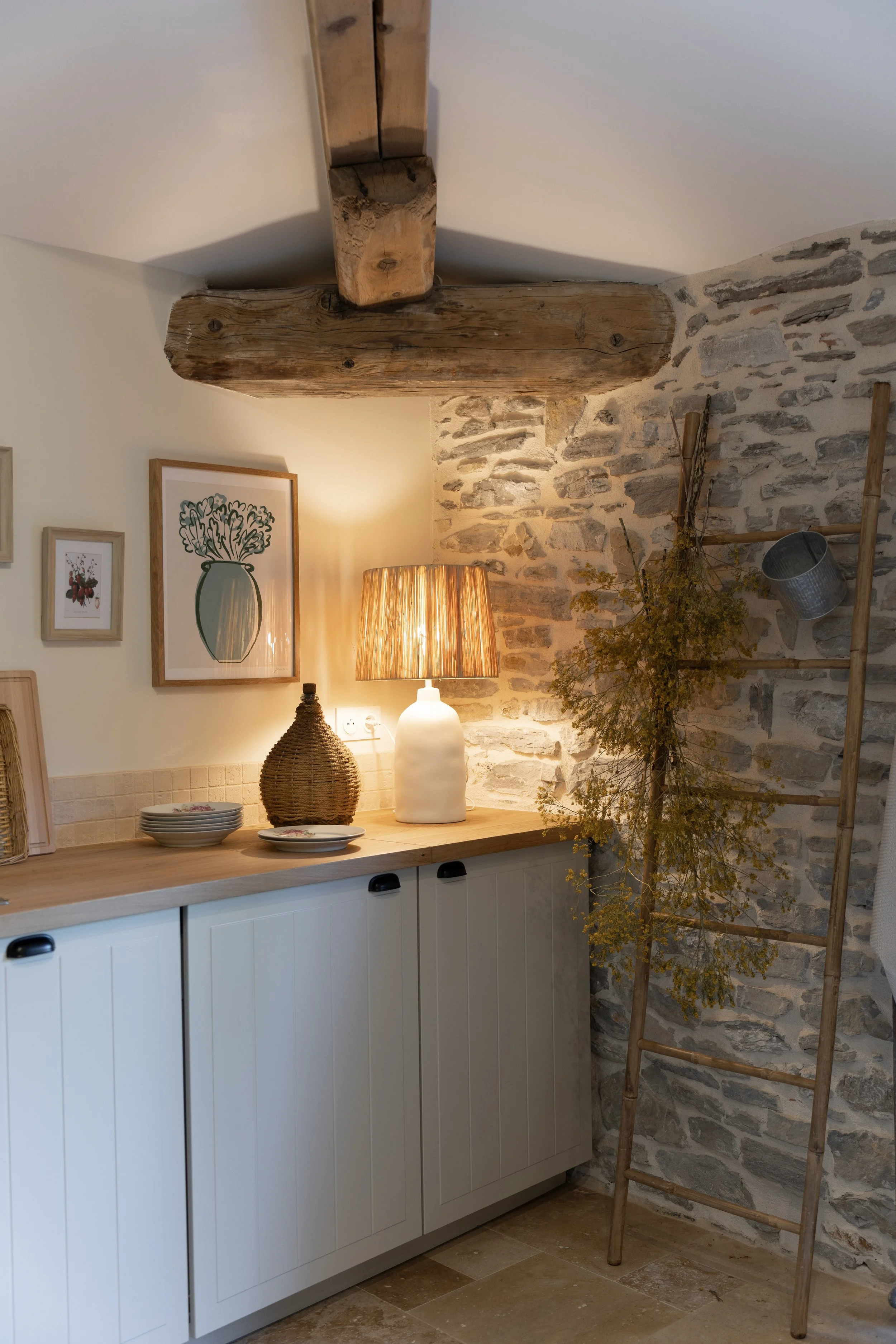 Intérieur de cuisine avec lampe sur table, cadres décoratifs, étagère en métal, branche sèche, et poutre en bois apparente au plafond.