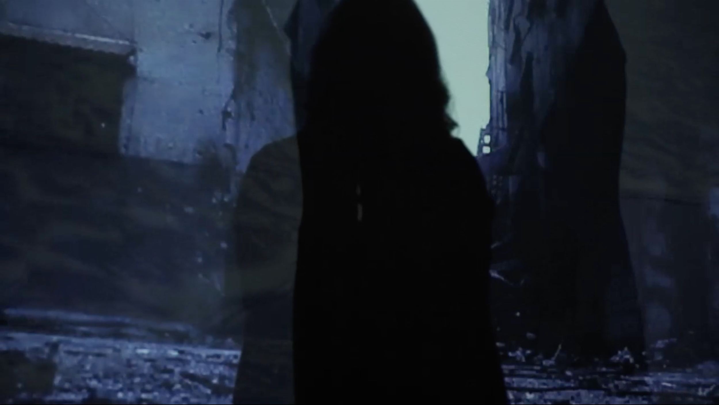 A person with long hair is looking into a dark, shadowy, narrow passage or alleyway with stone or concrete walls and a bright opening at the other end.