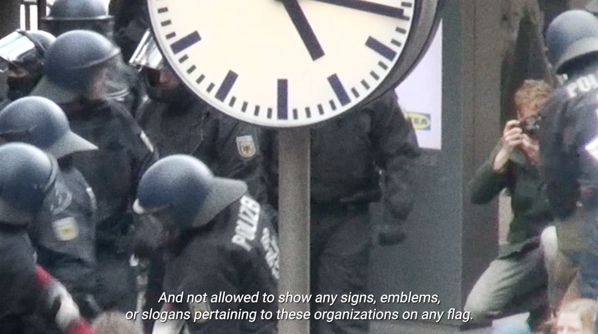 A group of police officers in uniform wearing helmets, standing near a large clock and a person taking a photo. The image appears to be taken in an indoor setting, possibly a police station or security area. There is a subtitle stating, 'And not allowed to show any signs, emblems, or slogans pertaining to these organizations on any flag.'