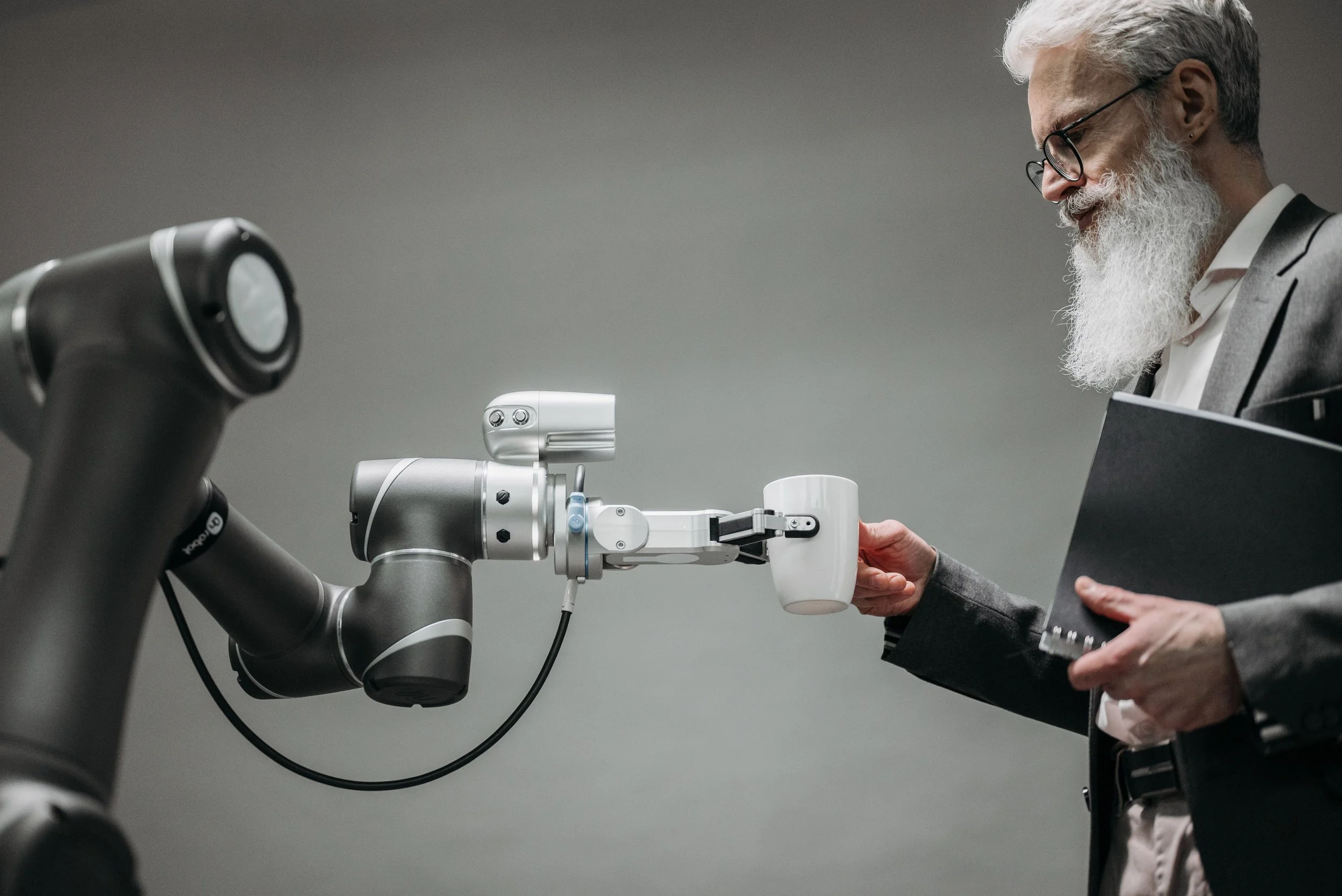 An elderly man with white hair and a long beard, wearing glasses and a grey blazer, is working with a robotic arm. He holds a white cup in one hand and a black notebook in the other, looking at the robotic arm.