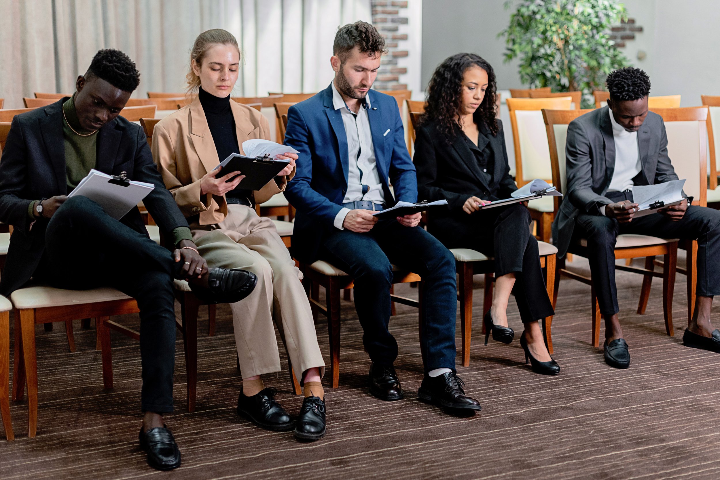 Five diverse business professionals sitting in a row, reading and reviewing documents during a meeting or seminar in a conference room.