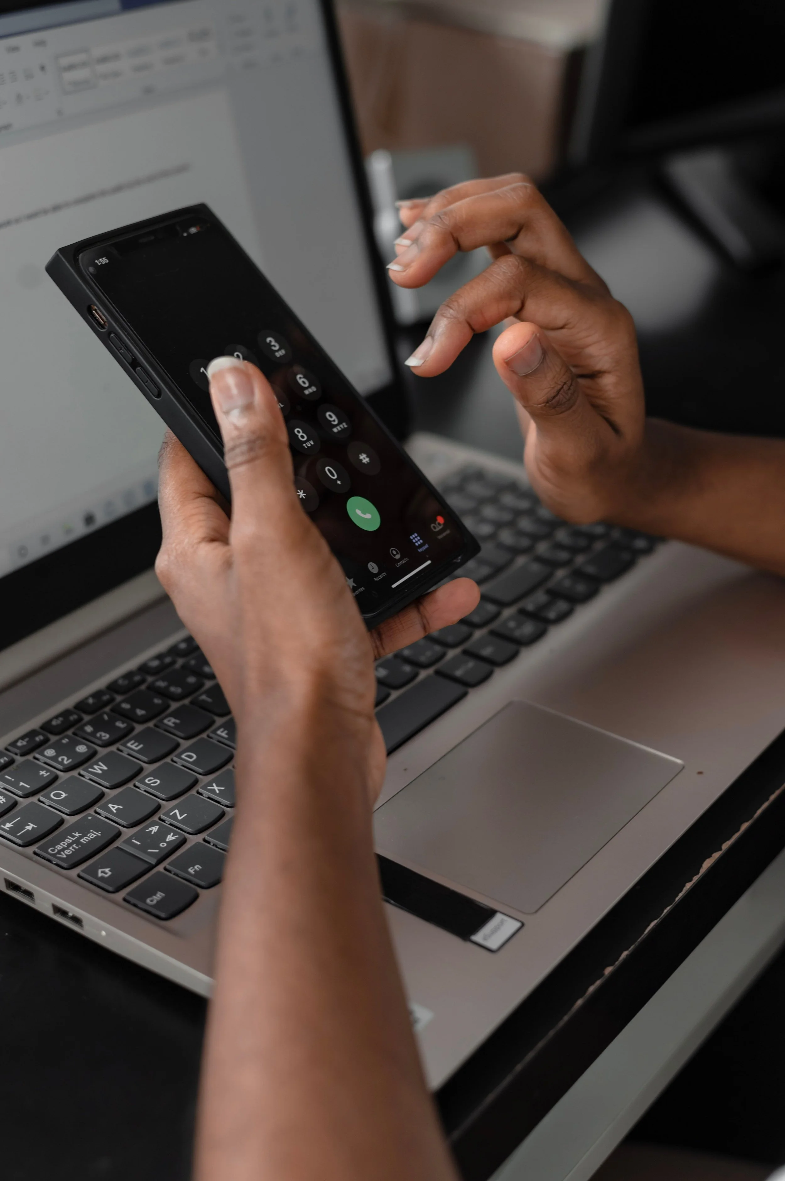 Close-up of a person's hands dialing a phone number on a smartphone held in their right hand, with a laptop keyboard partially visible in the foreground.