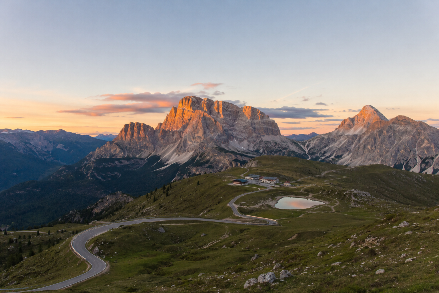Paisaje montañoso con picos rocosos, praderas verdes, una pequeña laguna y un camino sinuoso durante el atardecer.