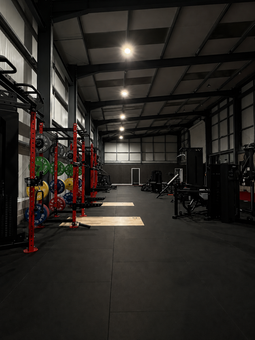 Empty gym with weight racks, barbells, and exercise equipment, in a spacious industrial-style interior with high ceilings and bright overhead lights.