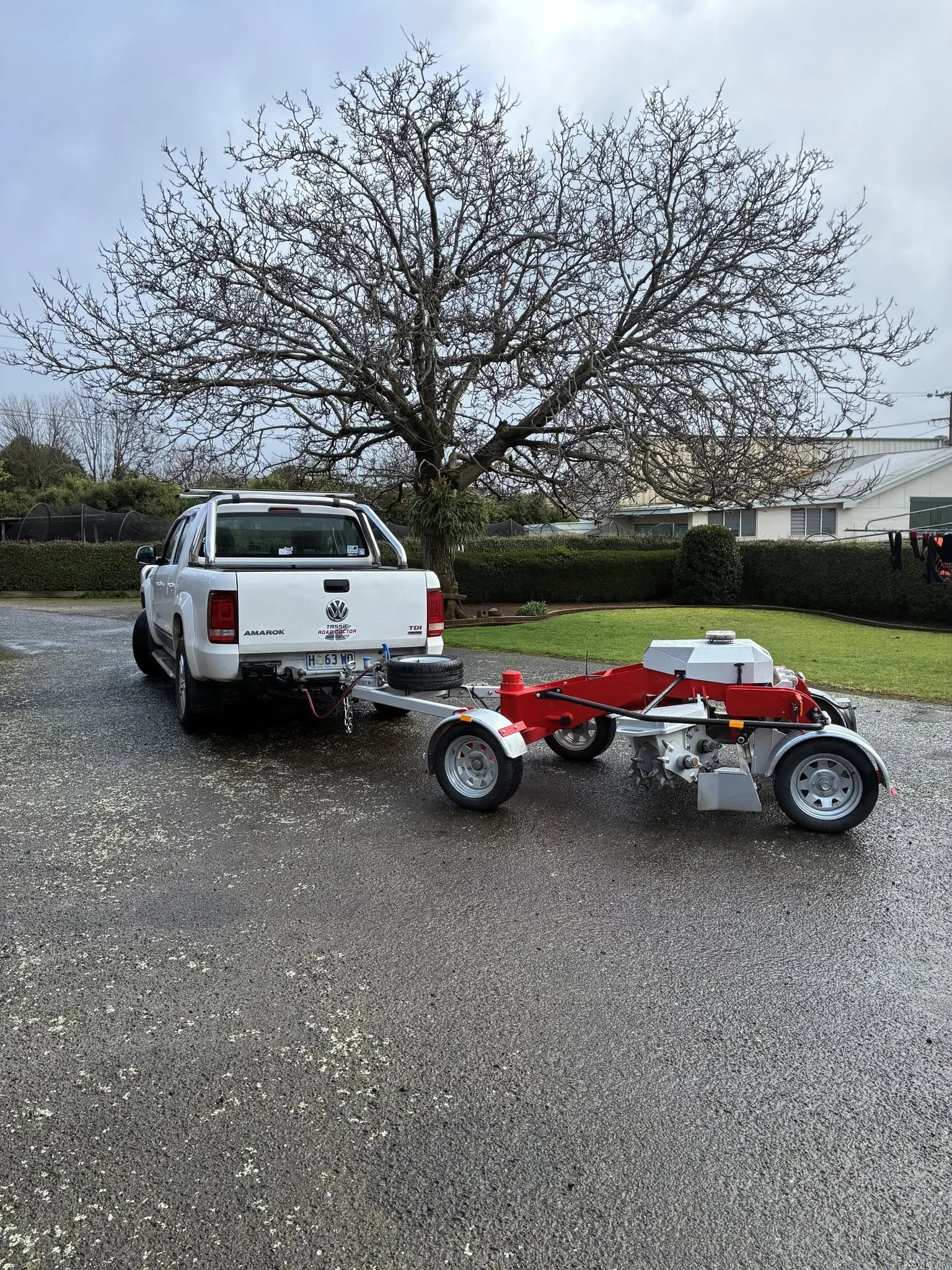 A white Volkswagen Amarok truck parked outdoors on a gravel surface, with a large leafless tree in the background and some green bushes. There is a red and white machine or equipment in the foreground.