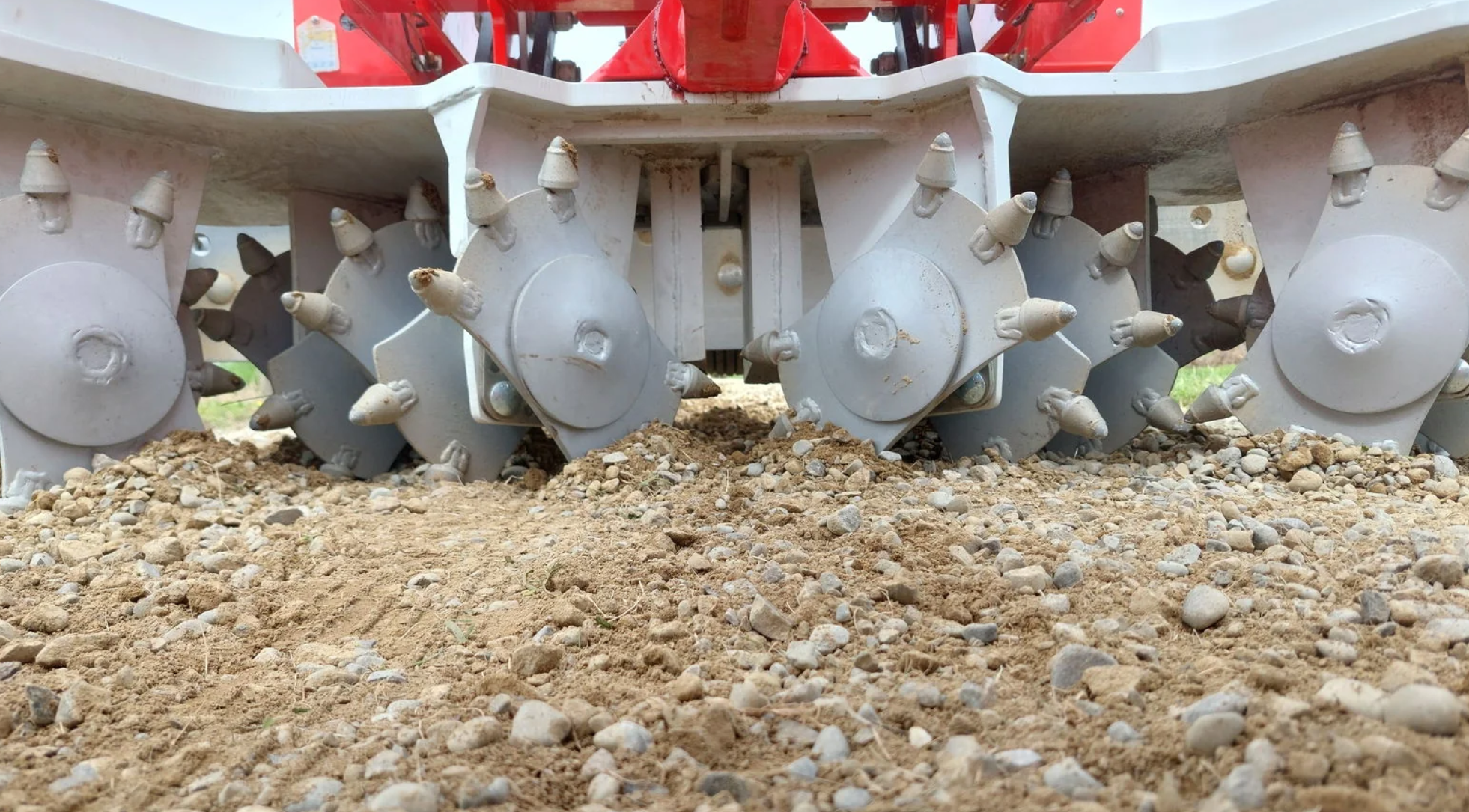 Close-up view of a tractor's tiller attachment with metal blades and spikes, working on a dirt and gravel surface.