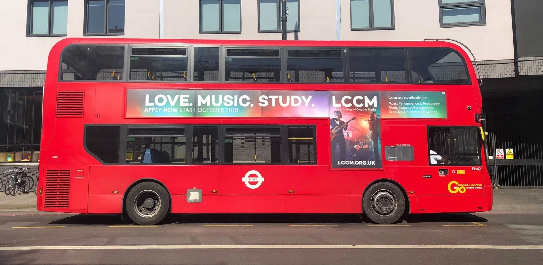 Red double-decker bus parked on the street with a large advertisement for London College of Creative Media and courses in music. Buildings in the background and bicycles parked on the sidewalk.