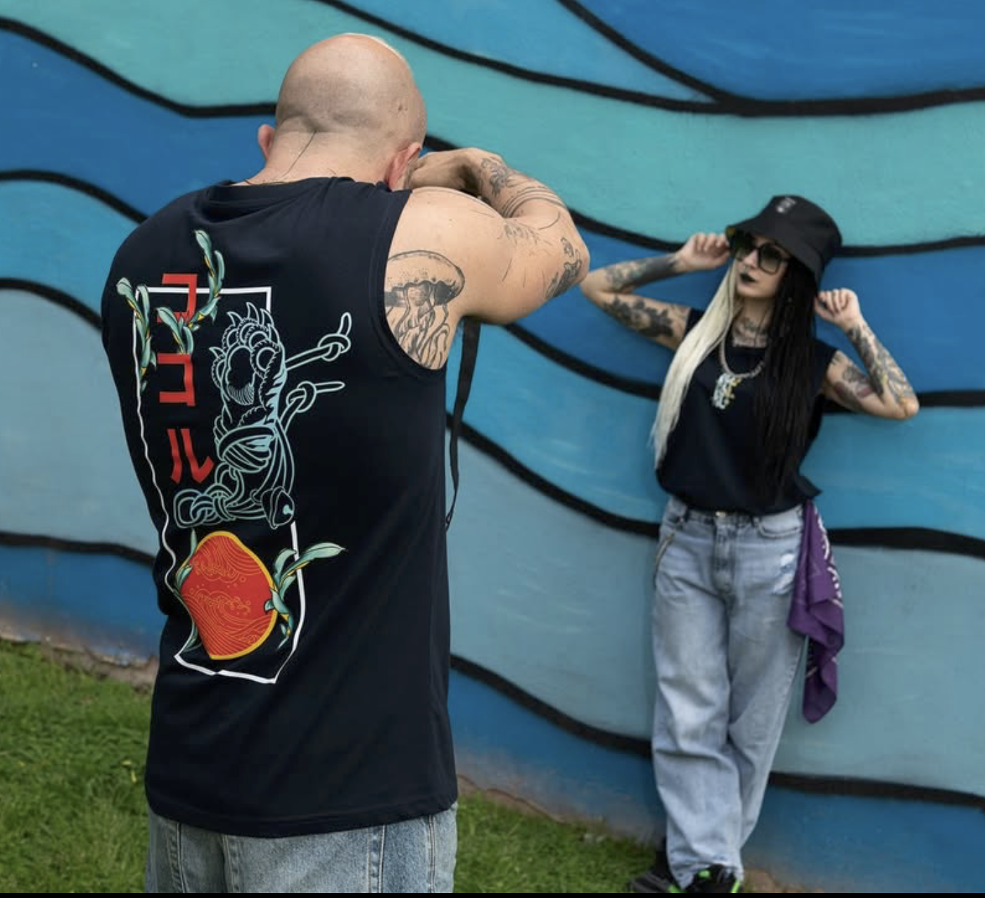 A man with tattoos is taking a photo of a woman who is standing against a colorful mural, wearing sunglasses, a black hat, and casual clothes.