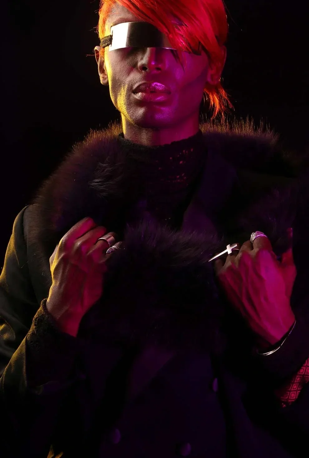 A person with short, red hair wearing a futuristic black visor, a black coat with fur collar, and jewelry, illuminated by purple and yellow lighting against a dark background.