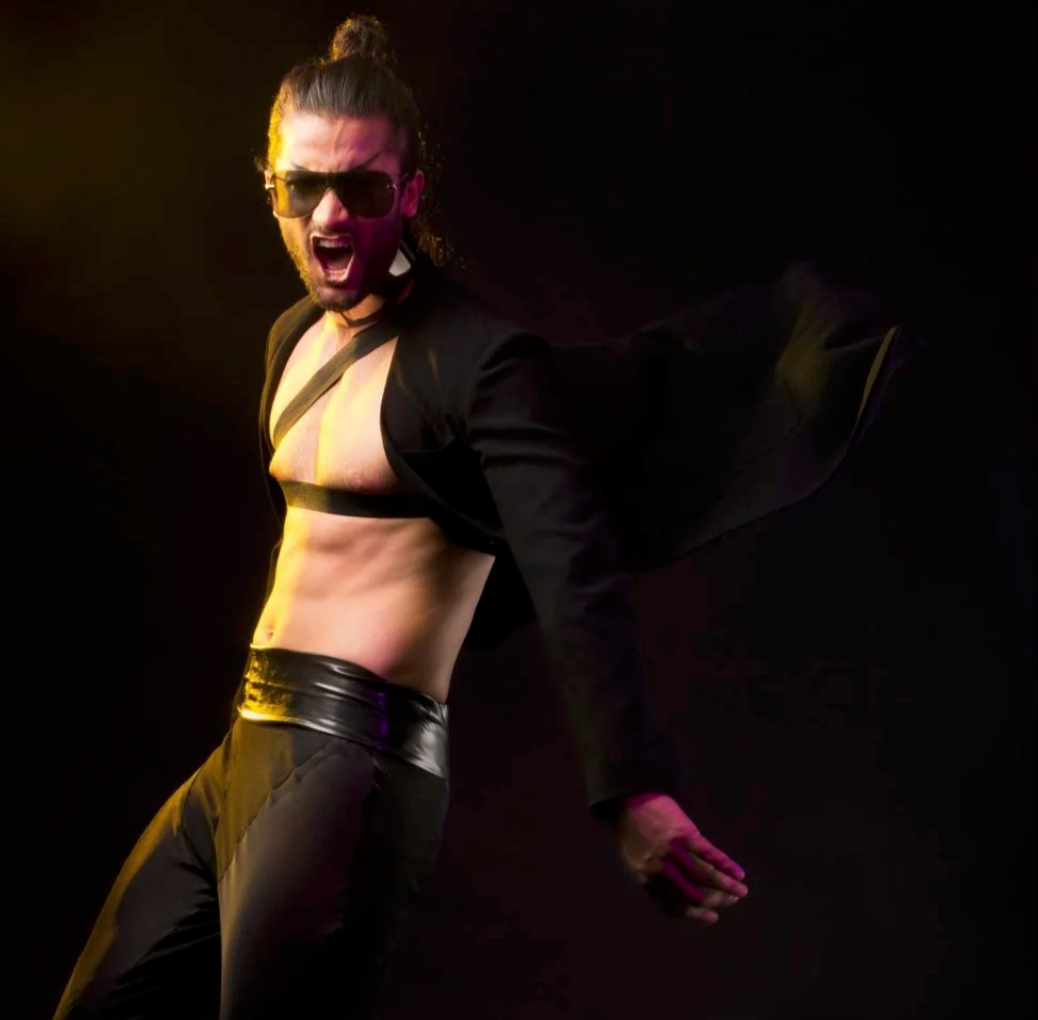 A man with long hair, beard, and sunglasses wearing black pants and an open black jacket, exposing his chest, striking a dynamic pose against a dark background with yellow and purple lighting.