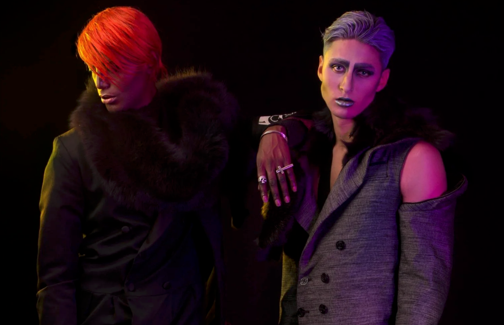 Two androgynous models in stylish clothing against a dark background with purple and yellow lighting.