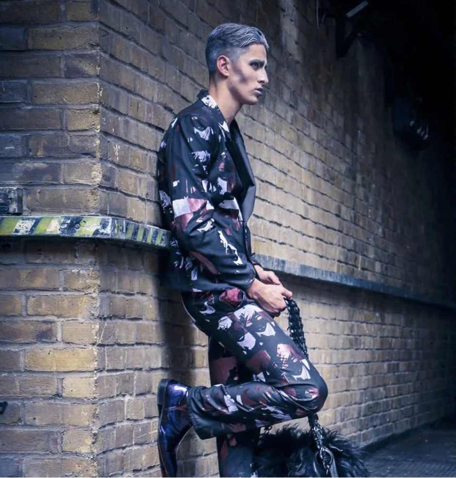 A woman leaning against a brick wall with short gray hair, wearing a matching dark, patterned jacket and pants, holding a leash with a black dog.