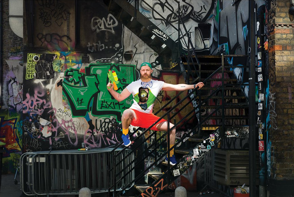 A man with a beard and colorful clothing poses on a graffiti-covered stairwell, holding a toy water gun, with graffiti art and tags on the walls.