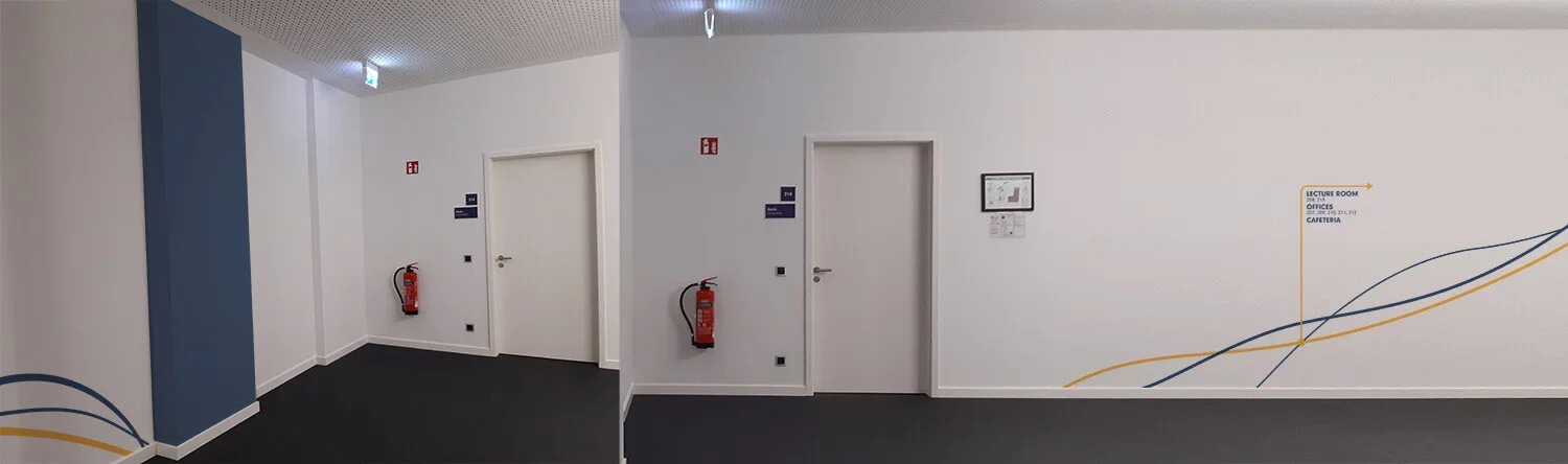 Interior of a building hallway with three doors, fire extinguishers, a wall-mounted display, and a sign indicating directions to lecture room, offices, and cafeteria.
