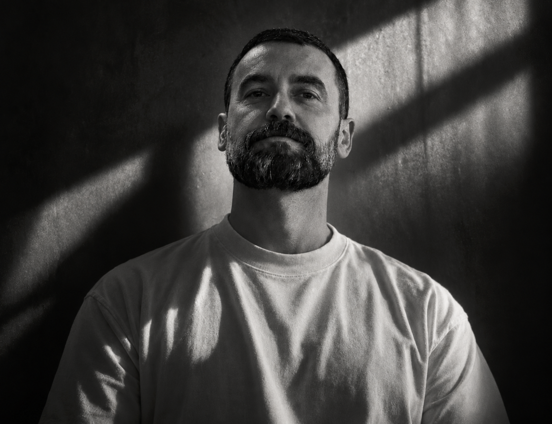 Black and white portrait of a man with dark hair, a beard, and a mustache, wearing a T-shirt, with dramatic shadowed lighting on the wall behind him.