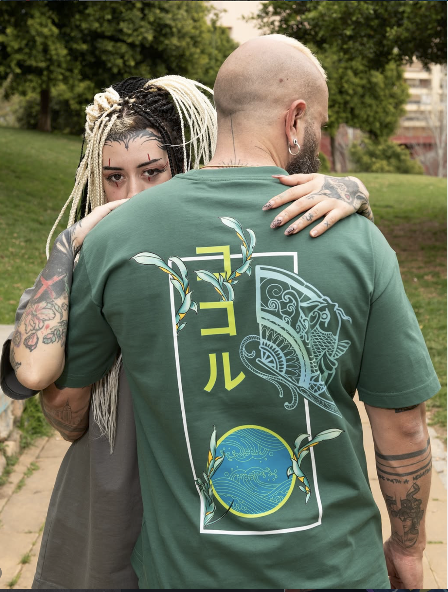 A woman with long braided hair and tattoos rests her head on a man's shoulder while hugging him from behind in a park. The man has a shaved head, tattoos, and is wearing a green T-shirt with a graphic design.