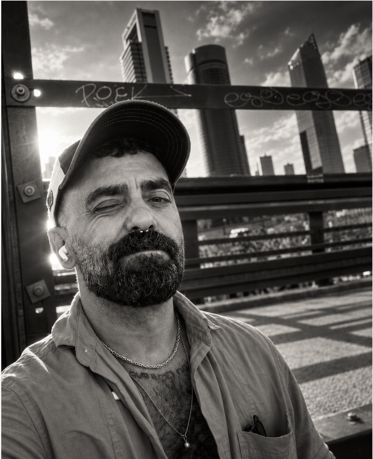 A man with a beard, mustache, and septum piercing winks at the camera in an urban setting with high-rise buildings in the background. He is wearing a baseball cap, a chain necklace, and a button-up shirt.
