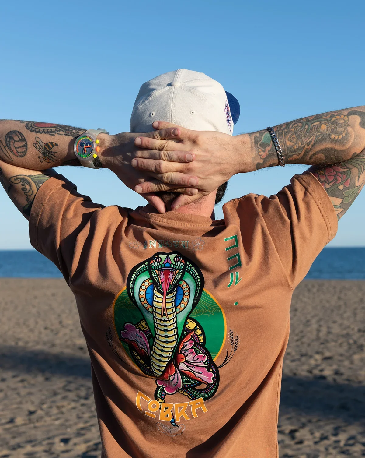 A person with tattoos on arms, wearing a brown T-shirt with a colorful cobra design, and a white cap, standing on a sandy beach with the ocean in the background, facing away from the camera with hands behind head.