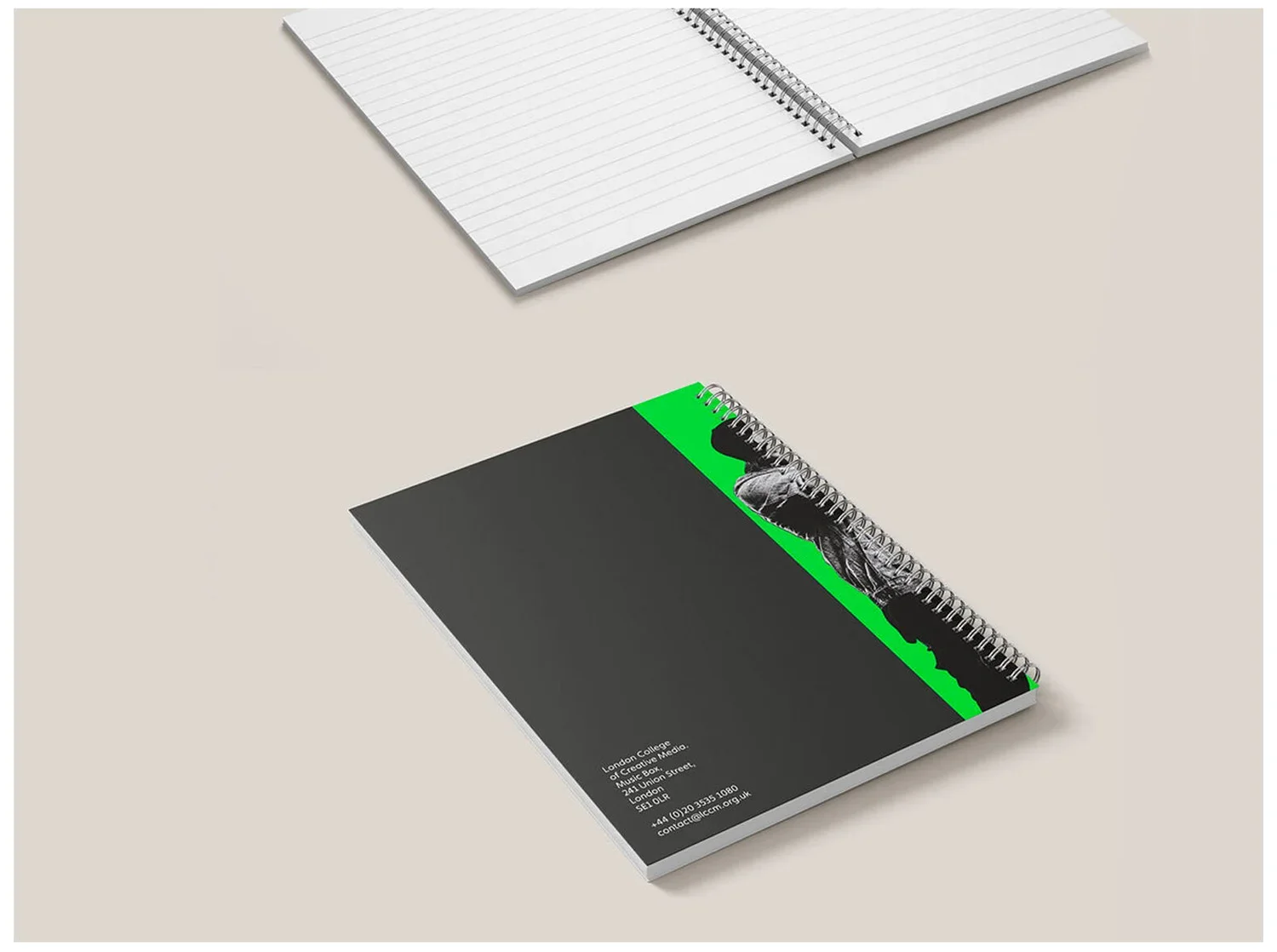 Two spiral notebooks placed on a light beige surface. The notebook in the foreground has a black cover with a green stripe and a black-and-white image of a person, and contact information at the bottom. The notebook in the background has a white cove
