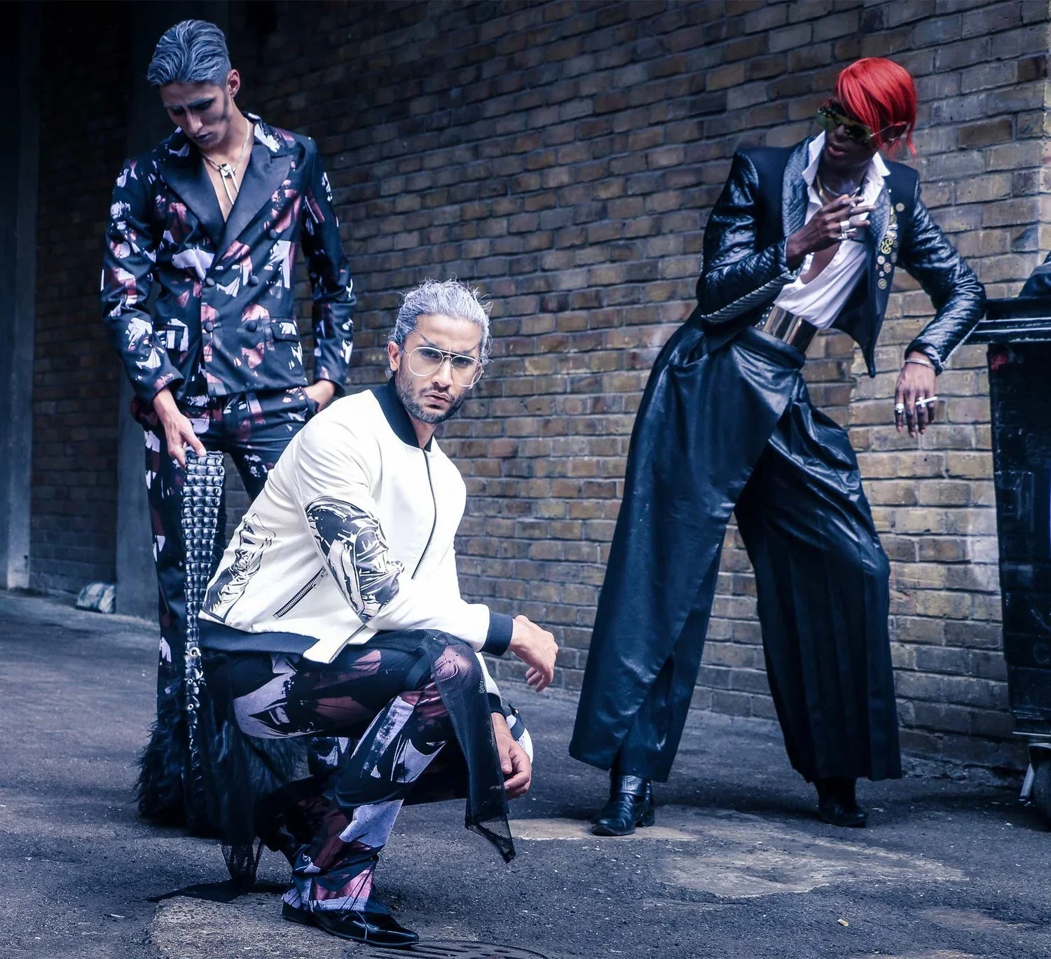 Four individuals dressed in fashionable clothing pose against a brick wall in an urban alleyway, with one crouching, one sitting, and two standing.