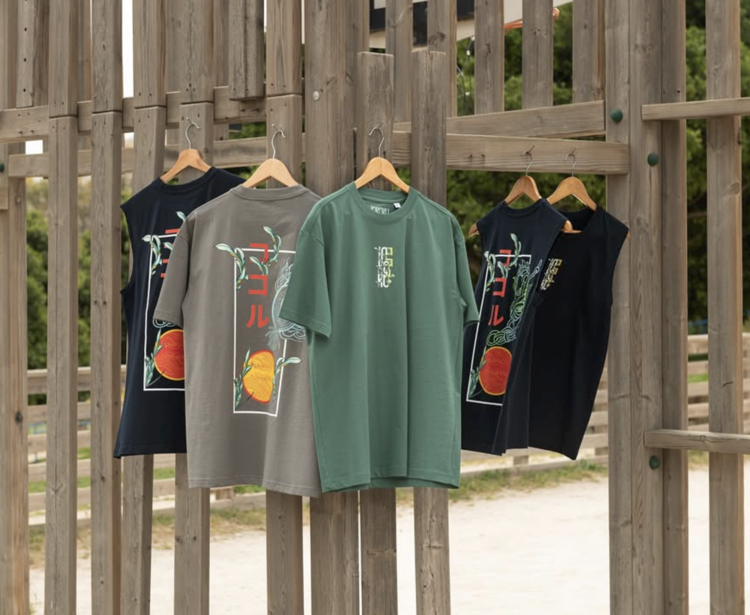 Five t-shirts hanging on wooden hangers on a wooden fence, with trees and a fence in the background. The t-shirts feature colorful graphics and Japanese characters.