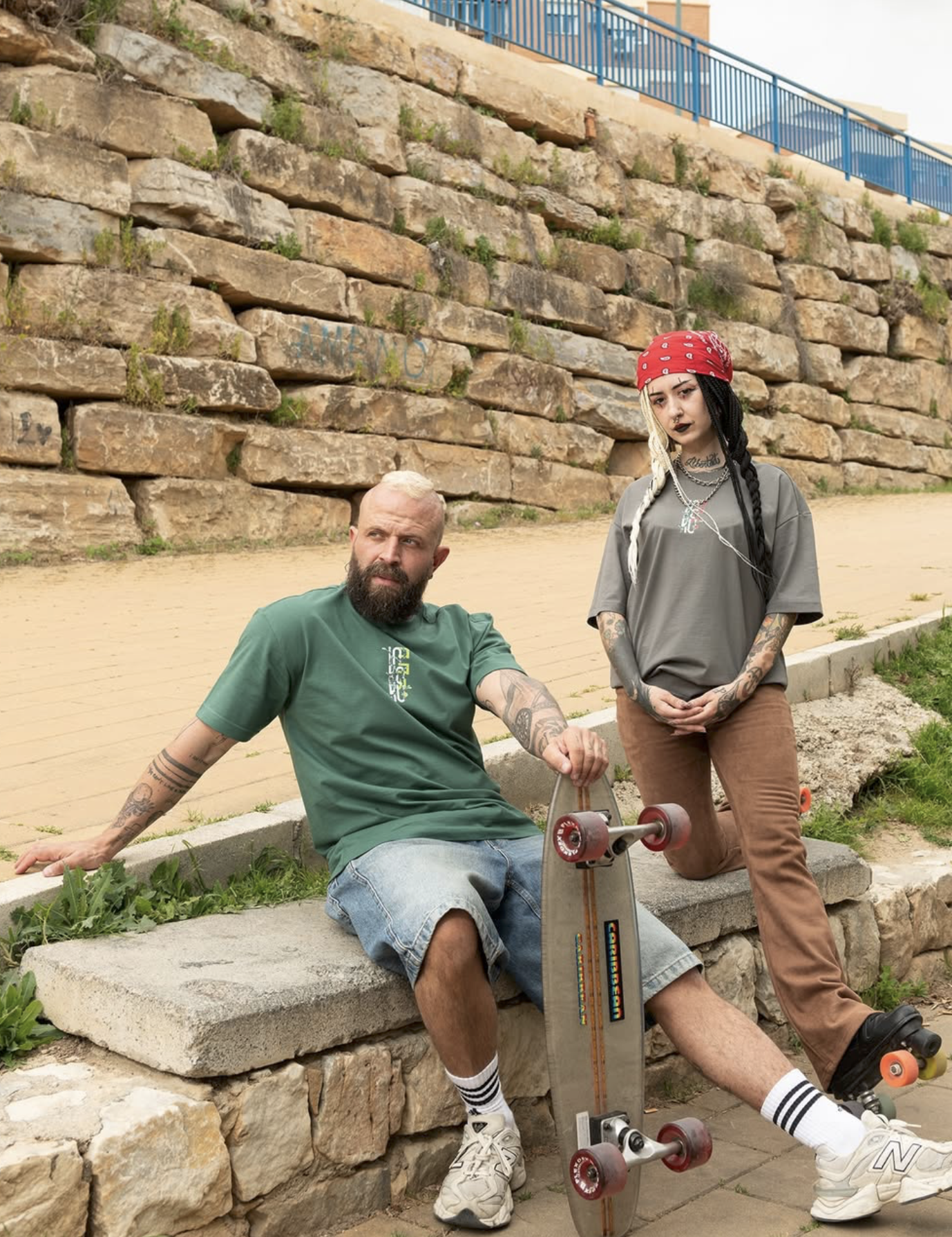 Two young adults with tattoos and piercings posing outdoors in front of a stone wall. One is sitting on the ground with a skateboard, wearing a green t-shirt and denim shorts. The other is standing, wearing a gray t-shirt, brown pants, a red bandana,