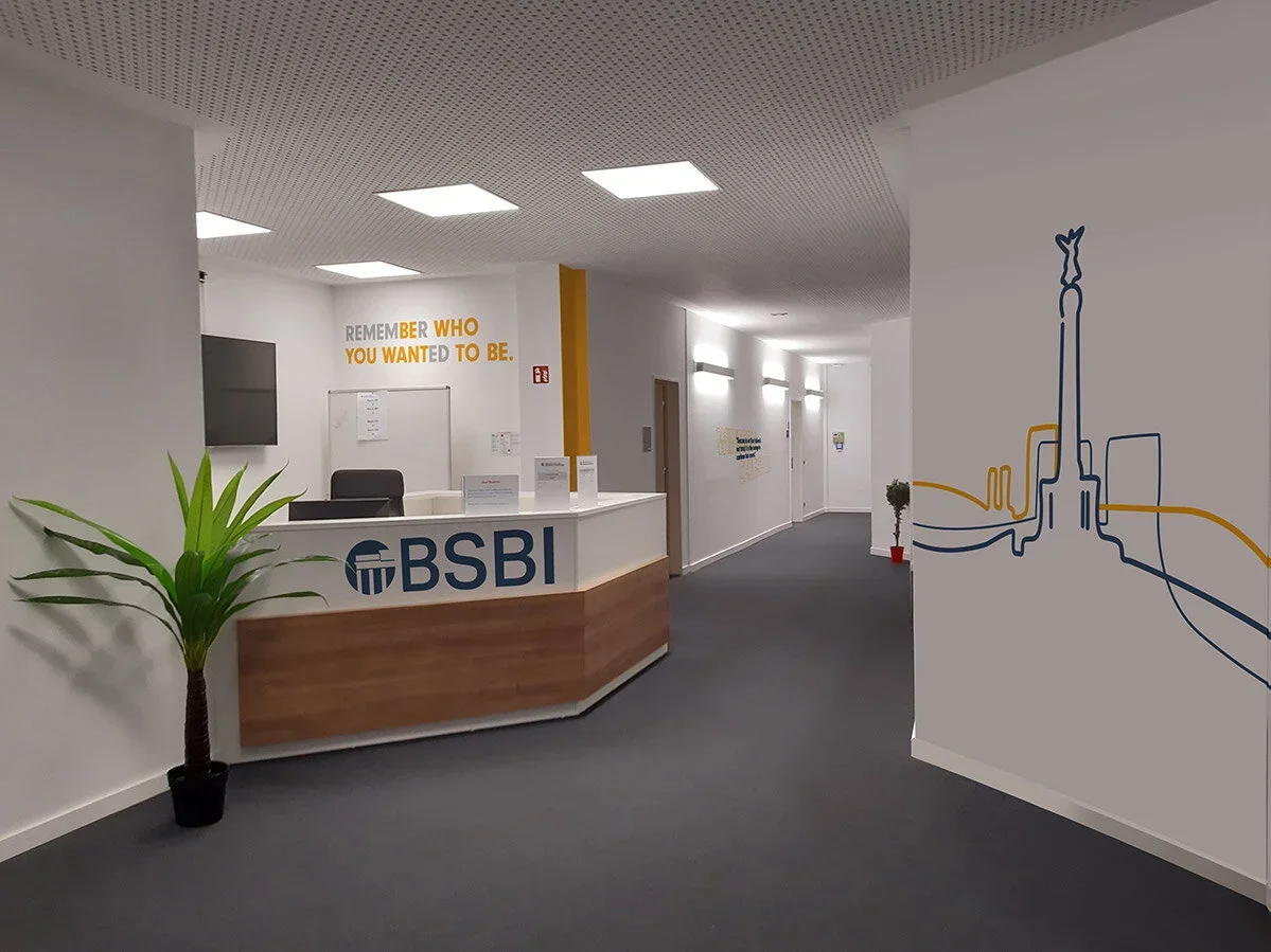 Office reception area with a BSBI logo on the front desk, a potted plant, wall art, and hallway in the background.