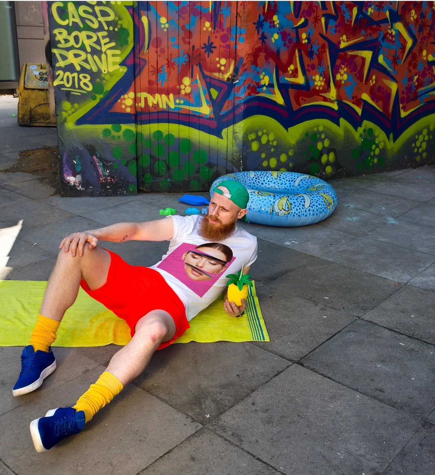 Two young men lying on the sidewalk in front of a colorful graffiti mural, one man is holding a pineapple and the other has a printed face mask on. They are dressed casually, with the man in the foreground wearing a white T-shirt, red shorts, yellow 