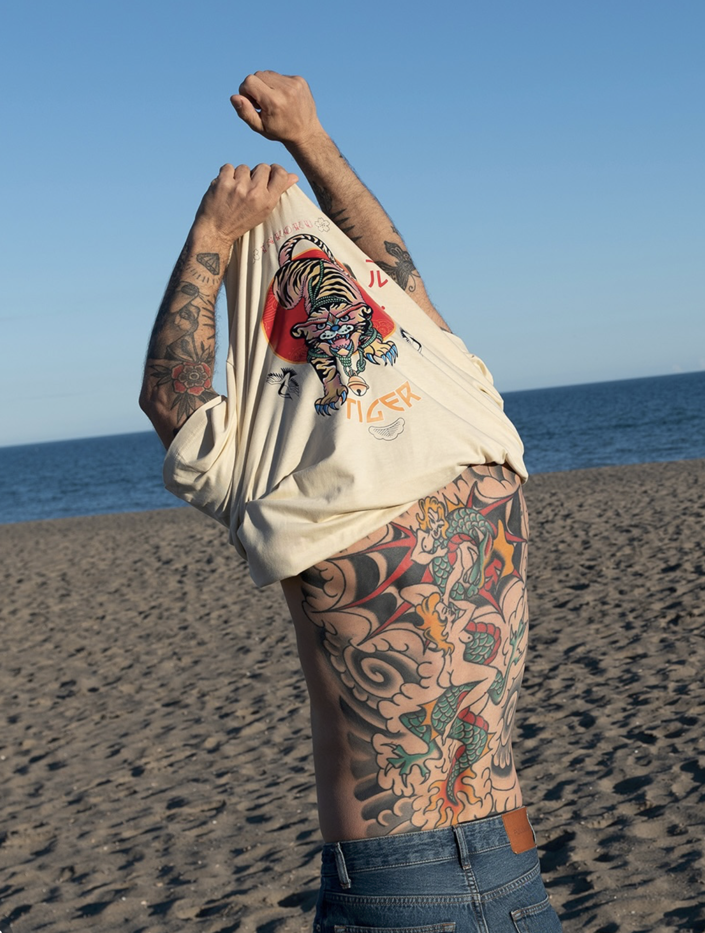 A person with tattoos on their arms and torso adjusting their shirt, standing on a sandy beach with the ocean and a clear blue sky in the background.