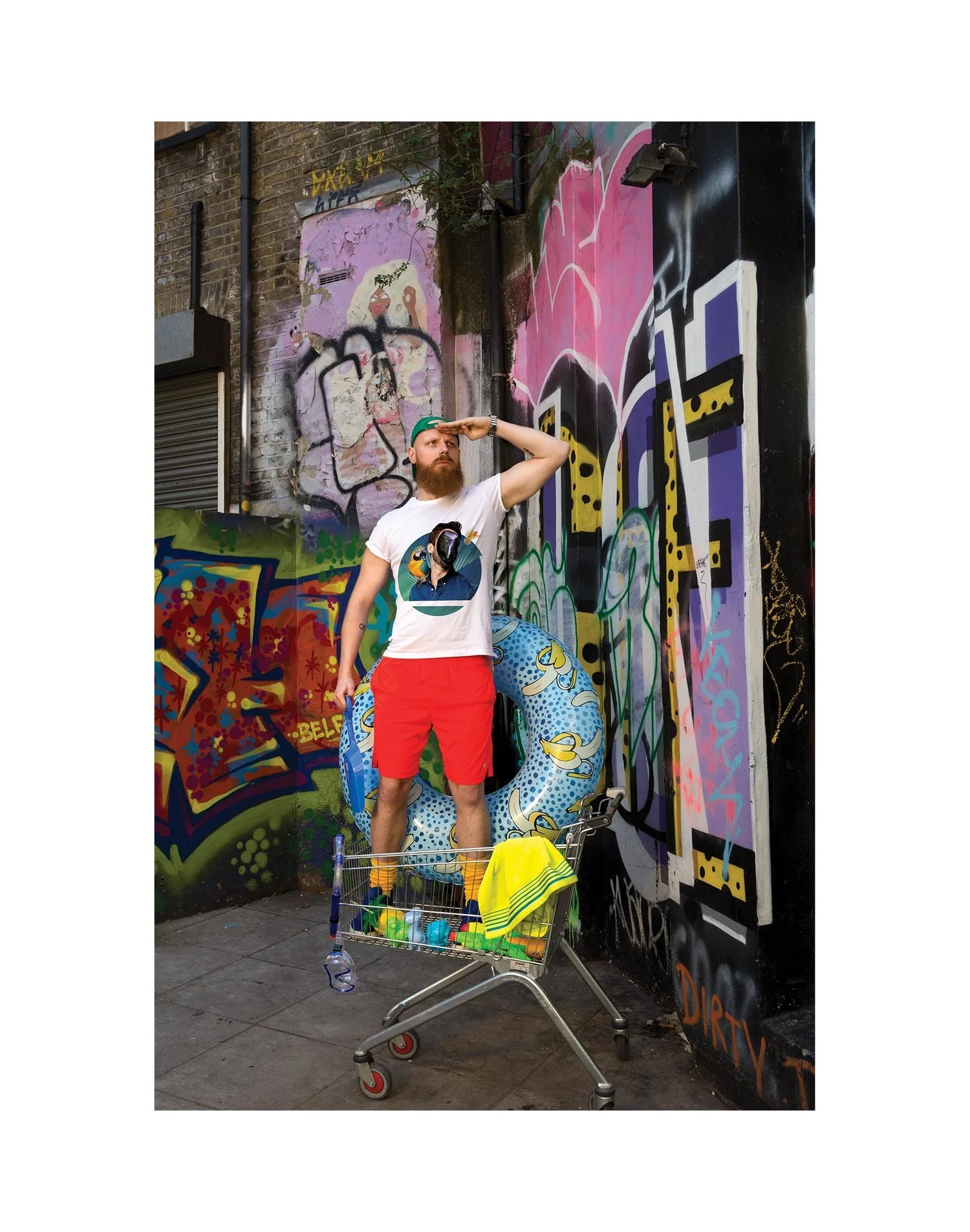 A man with a beard wearing a white t-shirt with a graphic of a woman, red shorts, and a green cap, stands in front of a graffiti mural on a brick wall. He is holding a peace sign with his right hand above his forehead and standing inside a pool float