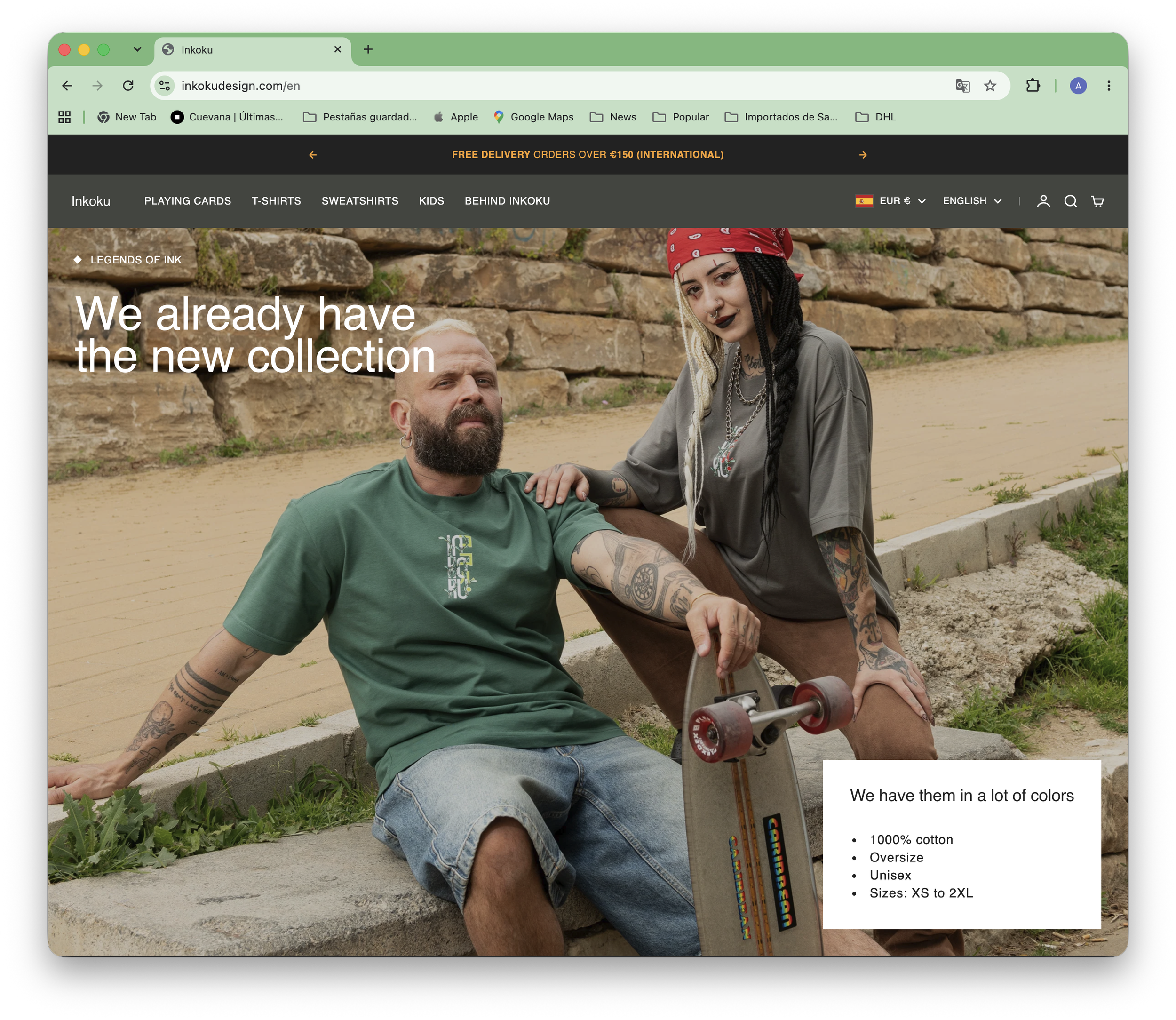 Two people sitting outdoors: one man with a beard and tattoos wearing a green t-shirt, and a woman with dreadlocks and tattoos wearing a gray t-shirt, sitting next to a skateboard. They are in a park or recreational area with a stone wall and greenery in the background.