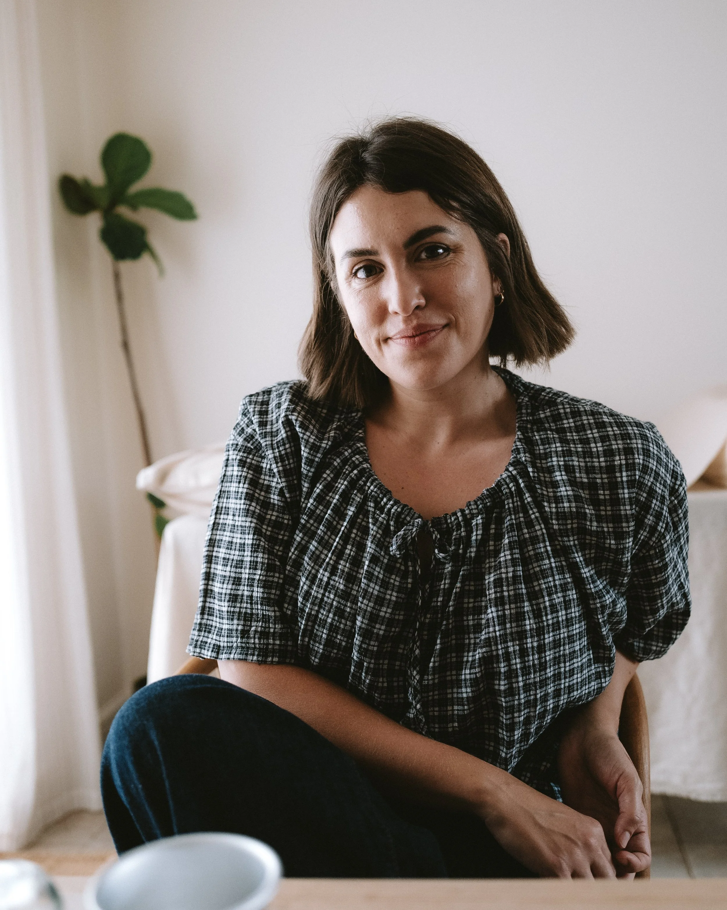 A woman with short brown hair and light skin, wearing a checkered shirt, sitting indoors with her hands resting on her knee, smiling softly at the camera, in a room with a white wall and a plant in the background.