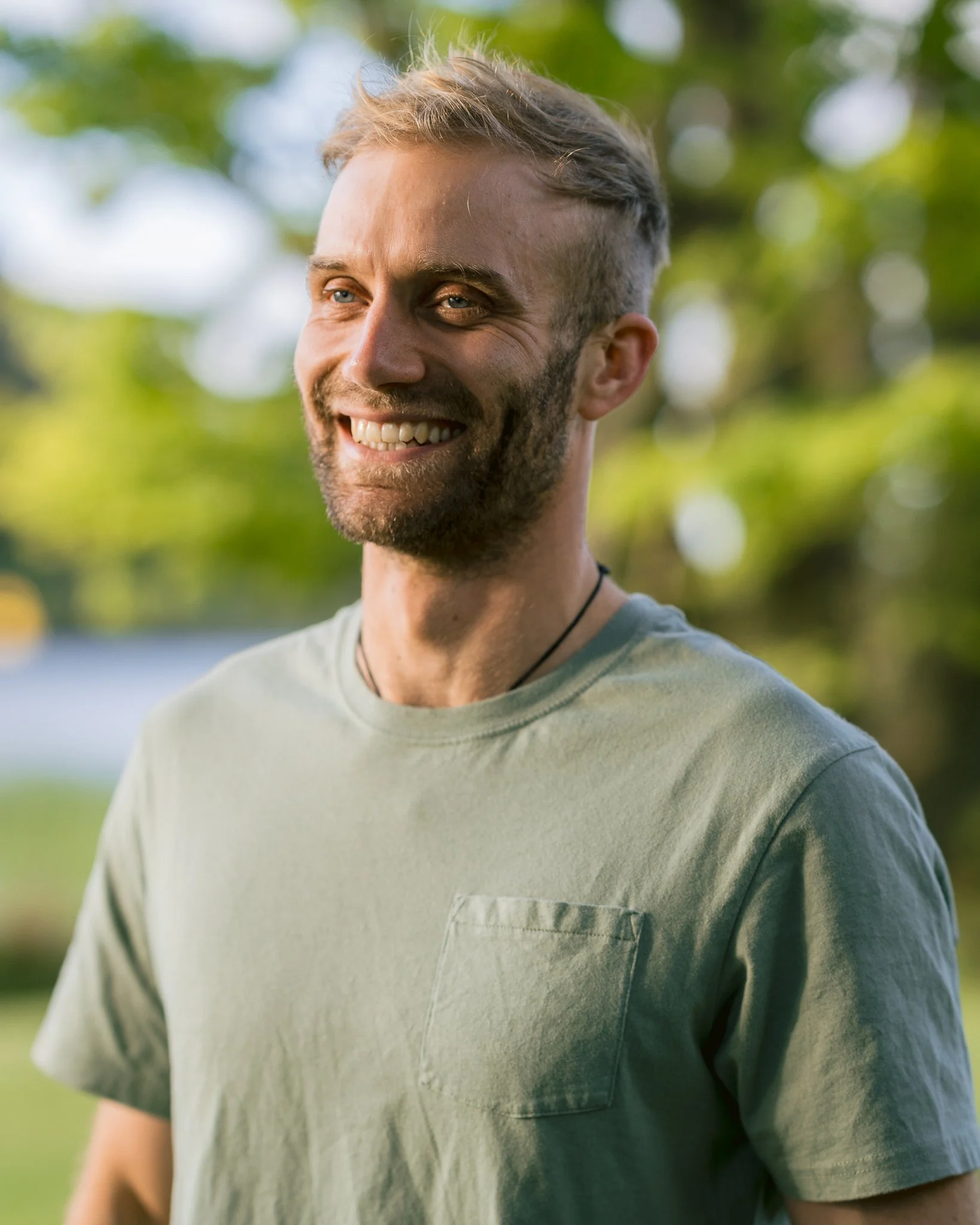 Meet Jesper: a professional, genuine and smiling counsellor in Papamoa and Tauranga with light blond hair and a dark beard, wearing a light-colored t-shirt, standing outdoors with a background of blurred green trees and sunlight.