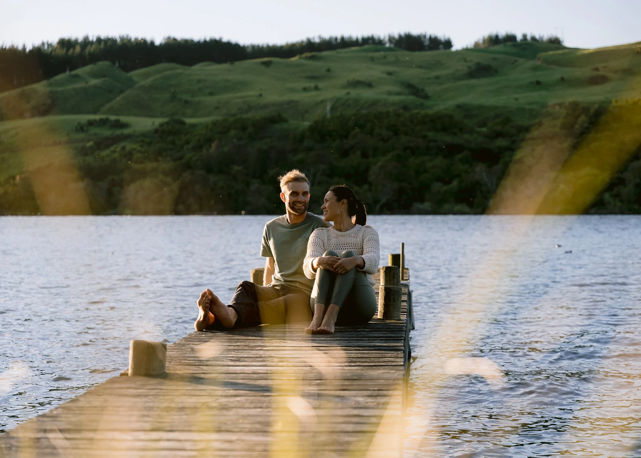 A couple sitting on a dock by a lake with hills and trees in the background during sunset, creating new positive emotional experiences together during couples counselling.