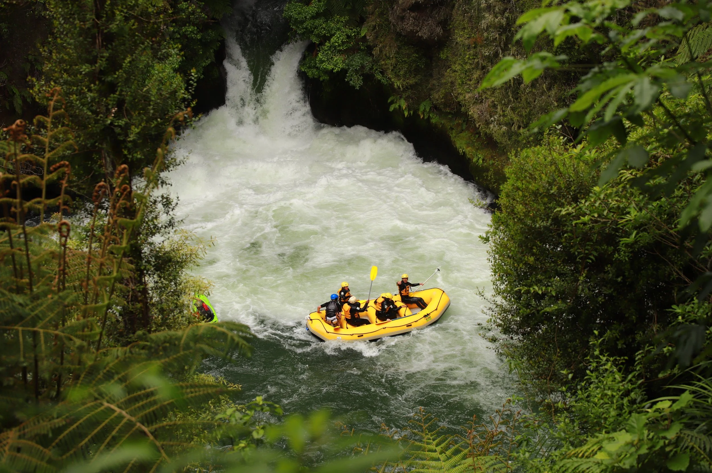 An adventure therapy group going white water rafting on the Kaituna River in the Bay of Plenty, surrounded by lush green trees and rocks, increasing confidence, resilience and connection with each other.