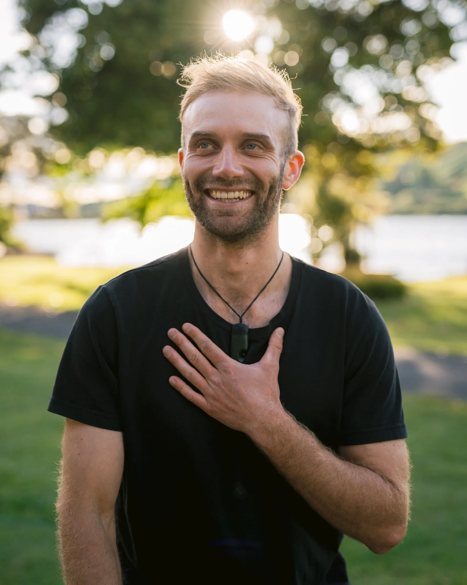 A young man with light-colored hair and a beard smiling outdoors, holding his hand over his chest, reconnecting with himself in Individual counselling, with the sun shining behind him and blurry trees and water in the background.