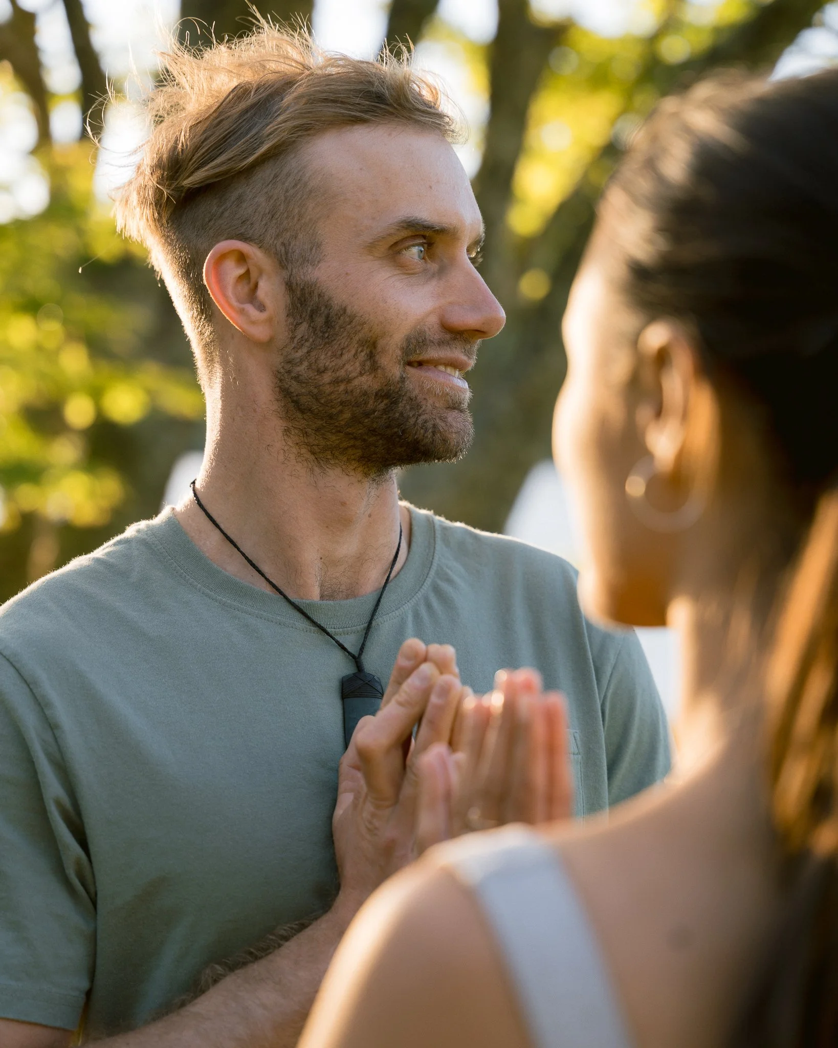 A young man with light skin, curly blonde hair on top, and a beard, looking at a woman with dark hair in an outdoor setting with trees and sunlight, creating a sense of space and connection during couples counselling.