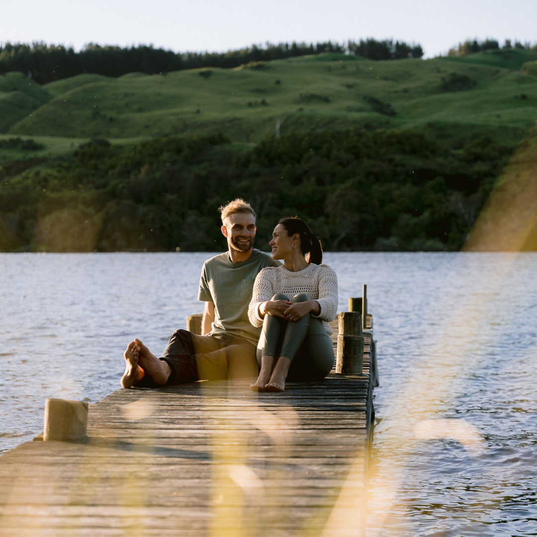 A couple sitting on a wooden dock by a lake, smiling and reconnecting with each other after couples counselling, with green hills in the background at sunset.